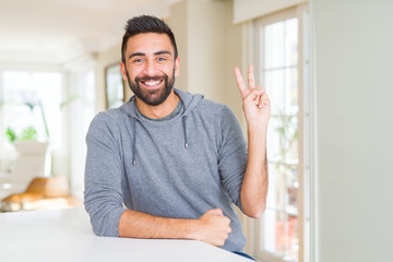 Handsome hispanic man wearing casual sweatshirt at home smiling with happy face winking at the camera doing victory sign. Number two.