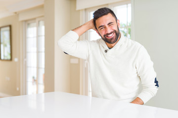 Handsome hispanic man wearing casual white sweater at home confuse and wonder about question. Uncertain with doubt, thinking with hand on head. Pensive concept.