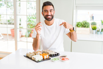 Handsome hispanic man eating asian sushi using chopsticks with surprise face pointing finger to himself