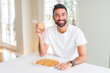 Handsome hispanic man eating healthy whole grain biscuits with a happy face standing and smiling...