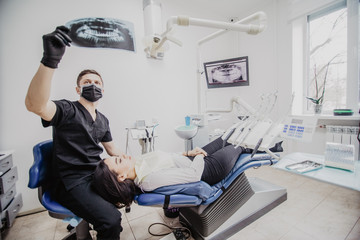 Young male dentist in dental office talking with female patient and preparing for treatment examining x-ray image.
