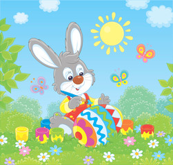 Little grey bunny coloring a big Easter egg on green grass among flowers on its front lawn near a small hut with thatched roof on a sunny spring day, vector illustration in a cartoon style