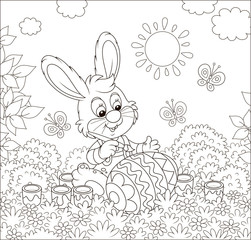 Little bunny coloring a big Easter egg on grass among flowers on its front lawn near a small hut with thatched roof on a sunny spring day, black and white vector illustration in a cartoon style