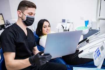Portrait of man dentist holding laptop while and demonstrate smth female patient at dental clinic