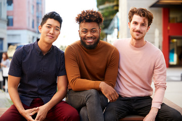 Three millennial male hipster friends on a bench in a city street smiling to camera, close up