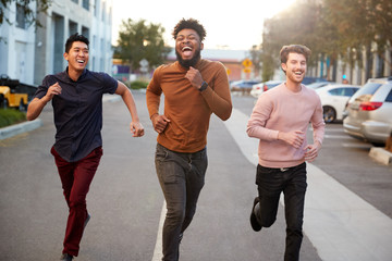 Three hip young adult male friends running for fun in a city road, front view, three quarter length