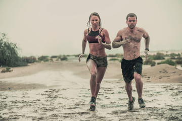 Muscular male and female athlete covered in mud running down a rough terrain with a desert background in an extreme sport race with grungy textured finish