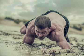 Closeup of strong athletic, muscular man crawling in wet muddy puddle in the rain in an extreme...
