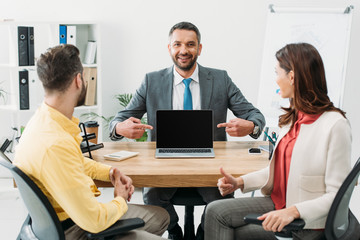 advisor pointing with fingers at laptop with blank screen near man and woman thumbing up in office