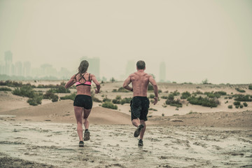 Rear view of muscular male and female athlete covered in mud running down a rough terrain with a desert background in an extreme sport race with grungy textured finish