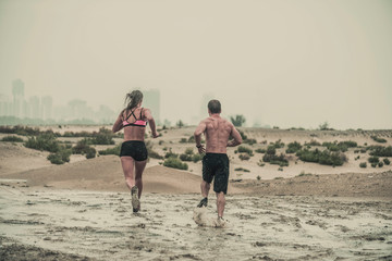 Rear view of muscular male and female athlete covered in mud running down a rough terrain with a desert background in an extreme sport race with grungy textured finish