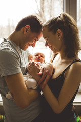 Woman and man holding on hands a newborn. On the background window. Mom, dad and baby. Portrait of young family. Happy family life. Man was born.