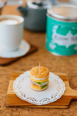 Mini Chicken Burger on wooden chopping board with blur tea cup and mug in the background.