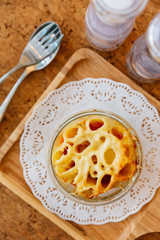 Top view of Cheese Baked Penne in glass bowl on wooden plate with salt, pepper and cutlery.
