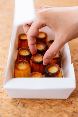 Female hand picking fresh baked Canelés inside white paper box. A small French pastry flavored with rum and vanilla with a soft and tender custard center and a dark, thick caramelized crust.