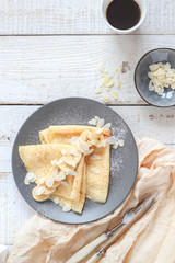 Crepes with bananas and almond slices and coffee on a white wooden table