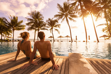 Couple enjoying beach vacation holidays at tropical resort with swimming pool and coconut palm trees near the coast with beautiful landscape at sunset, honeymoon destination - obrazy, fototapety, plakaty