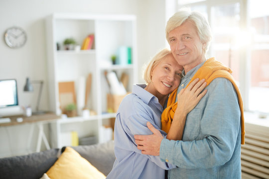 Waist up portrait of happy senior couple embracing tenderly at home and looking at camera