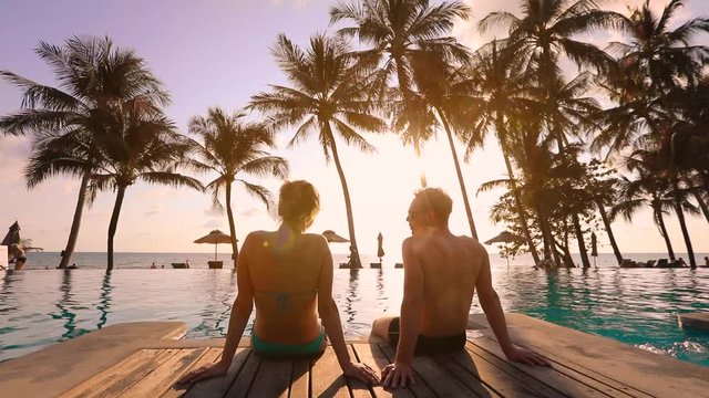 Couple relaxing at tropical beach resort and talking together at sunset, summer vacation holidays destination, scenic landscape with the sea and coconut palm trees, romantic honeymoon