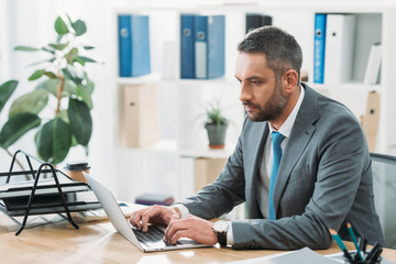 Naklejka premium handsome businessman sitting at table with laptop and typing in office