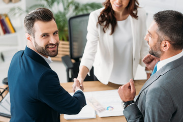 selective focus of advisor in suit and handsome investor shaking hands at workplace