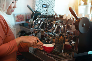 Female Muslim barista using espresso machine to pour coffee in cup at cafe