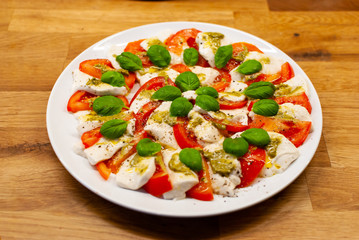 Angled view of plate full of vegan Mozzarella made of cashew nuts with tomato slices, basil and pesto on a wooden table