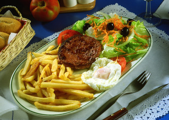 Plato con una ensalada de lechuga, tomate, aceitunas, zanahoria, un huevo frito, una hamburguesa y patatas fritas.
