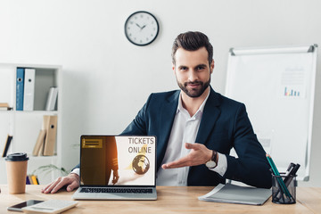 Naklejka premium handsome advisor in suit pointing with fingers at laptop with tickets online website on screen at office