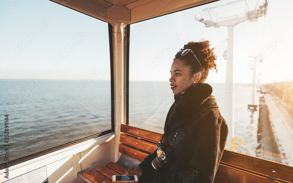 Wall mural a charming amazed african-american tourist girl in a coat is sitting inside of the cabin of a funicu