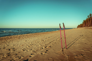 Vintage peaceful evening sunset Baltic sea pine tree dune coast beach with footprints and nordic walking poles close-up, high contrast blue cloudless sky and light waves in background
