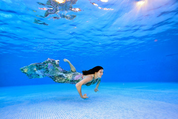 Girl swims in a dress underwater near the bottom of the pool on a blue background and smiles. Portrait. Shooting underwater. Landscape view