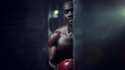 Fototapeta na wymiar Hands of boxer over dark gym background. Strength, attack and motion concept. Fit african american model in movement. Naked muscular athlete in red gloves. Sporty man during boxing
