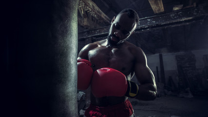 Hands of boxer over dark gym background. Strength, attack and motion concept. Fit african american model in movement. Naked muscular athlete in red gloves. Sporty man during boxing