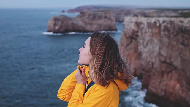 Close Up Portrait Of Young Woman Standing On The Wind, Stormy Ocean Background. SLOW MOTION. Breeze is playing with girls hair. Female traveler in yellow raincoat exploring Atlantic coast in Portugal.