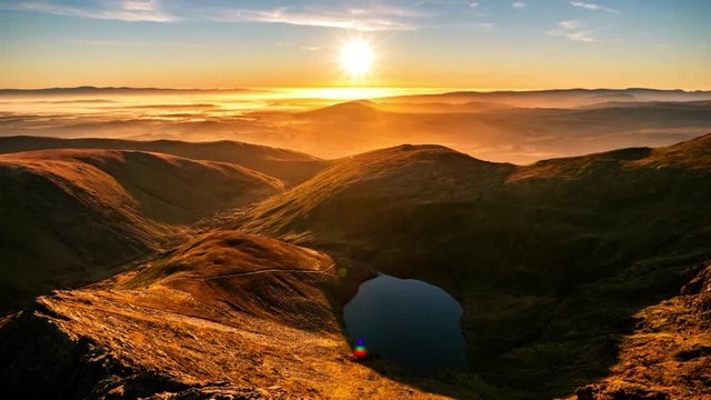 A time-lapse of a stunning orange yellow sunrise in Spring Summer from Sharp Edge on Blencathra, Lake District, Cumbria with reflection in lake tarn and cloud inversion over mountains in the distance