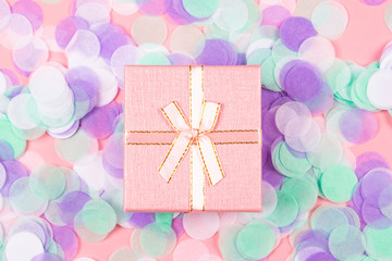 Little cute present box with shiny satin bow on pink background with multicolored confetti. Flat lay style. Holiday and celebration concept.