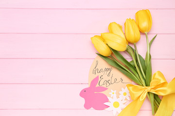 Yellow tulips with paper rabbit and text Happy Easter on pink wooden table