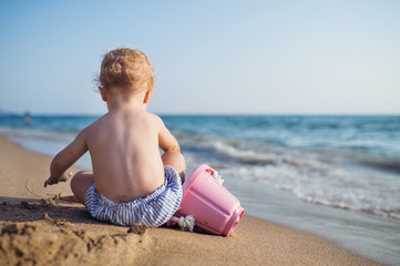A rear view of small toddler girl sitting on beach on summer holiday, playing.