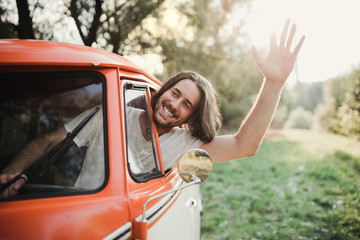 A young man driver in a car on a roadtrip through countryside, waving.