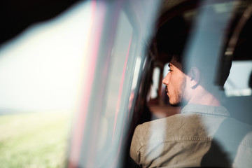 A young man sitting in a car on roadtrip through countryside, shot through glass.