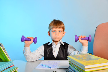 The little schoolboy is engaged in dumbbells among notebooks and textbooks at his desk at school in...