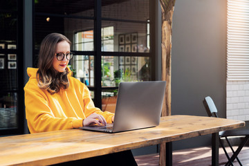 Front view. Girl in yellow hoodie sits in cafe in front of open laptop computer. Woman works on computer, checks email.