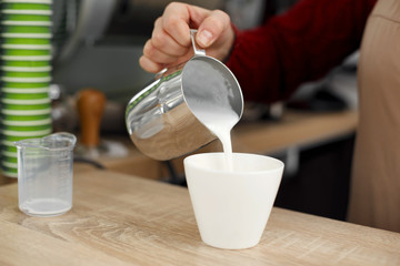 Barista pours milk from metal pot to white glass cup on wooden table.Prepait latte
