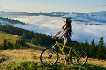 Fototapeta premium Young female rider standing with yellow bicycle in the mountains, wearing helmet, enjoying valley view on summer morning. Foggy mountains, forests on the blurred background. lifestyle concept