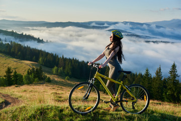 Young female rider standing with yellow bicycle in the mountains, wearing helmet, enjoying valley view on summer morning. Foggy mountains, forests on the blurred background. lifestyle concept