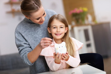 Young girl and her mother with piggybank sitting at table