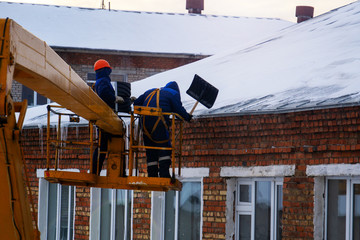  Workers in overalls and orange helmets on the crane basket remove icicles from roof of the house on a winter day - cleaning the roofing, utility service, safety, top view