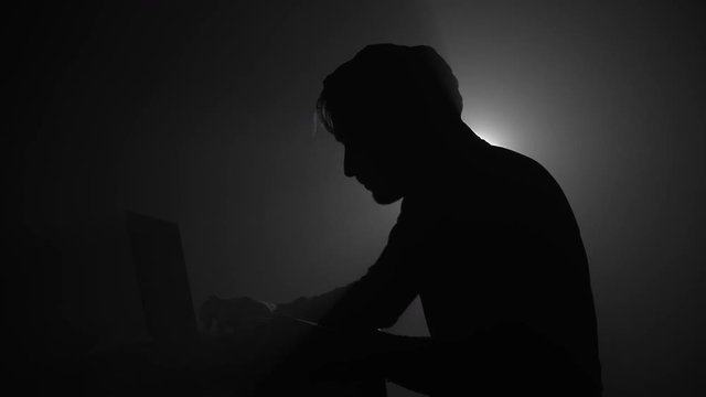 Silhouette of computer hacker in hoodie with laptop in dark