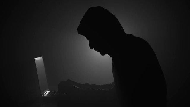 Silhouette of computer hacker in hoodie with laptop in dark